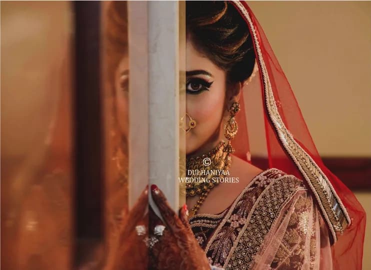 Bride in a red dupatta and embroidered lehenga peeking out from behind a door